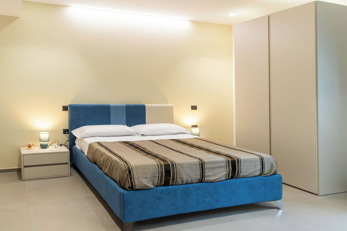 Ein modernes Schlafzimmer mit einem blauen Bett, weißen Kissen, einem Nachttisch mit Lampe und einem Kleiderschrank.