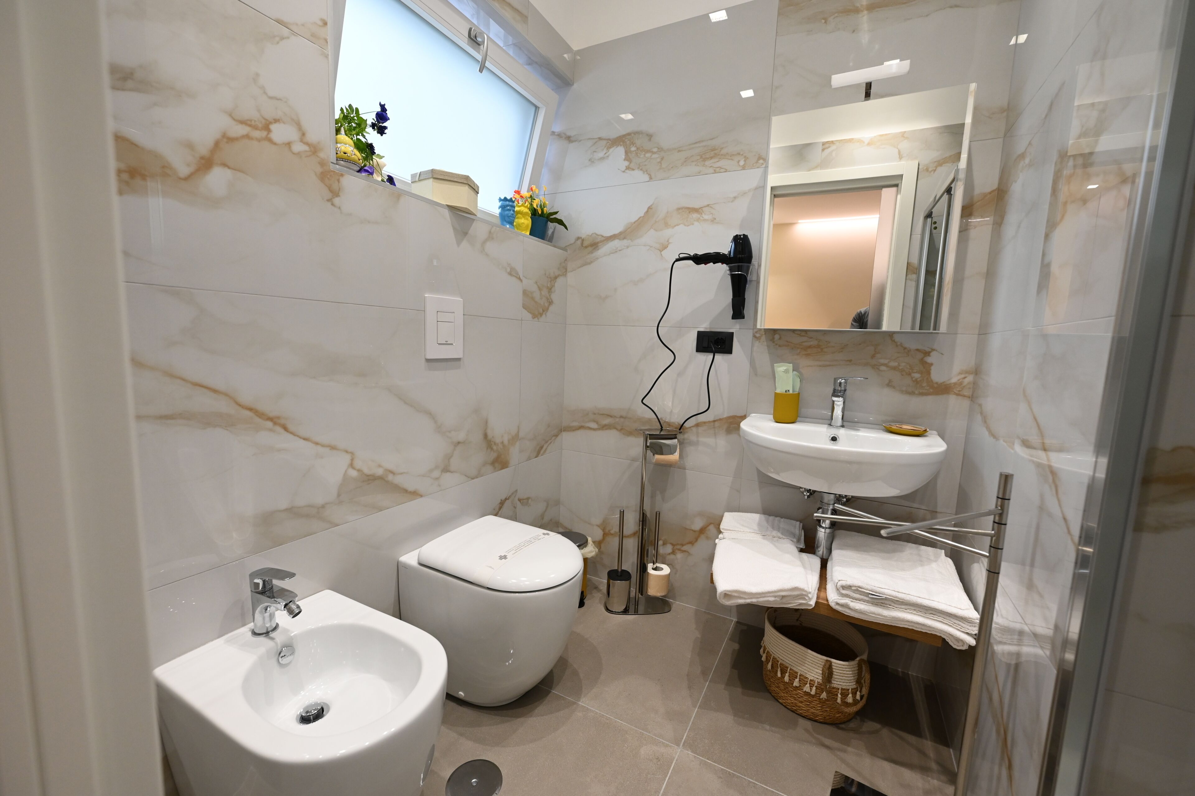 Uma casa de banho moderna com parede de mármore, lavatório, bidé, secador de cabelo e janela com vista.