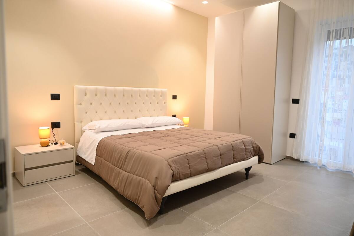 Ein modernes Schlafzimmer mit einem Bett, Nachttischlampen, einem Nachttisch, einem Kleiderschrank und einem Fenster mit durchsichtigen Vorhängen.