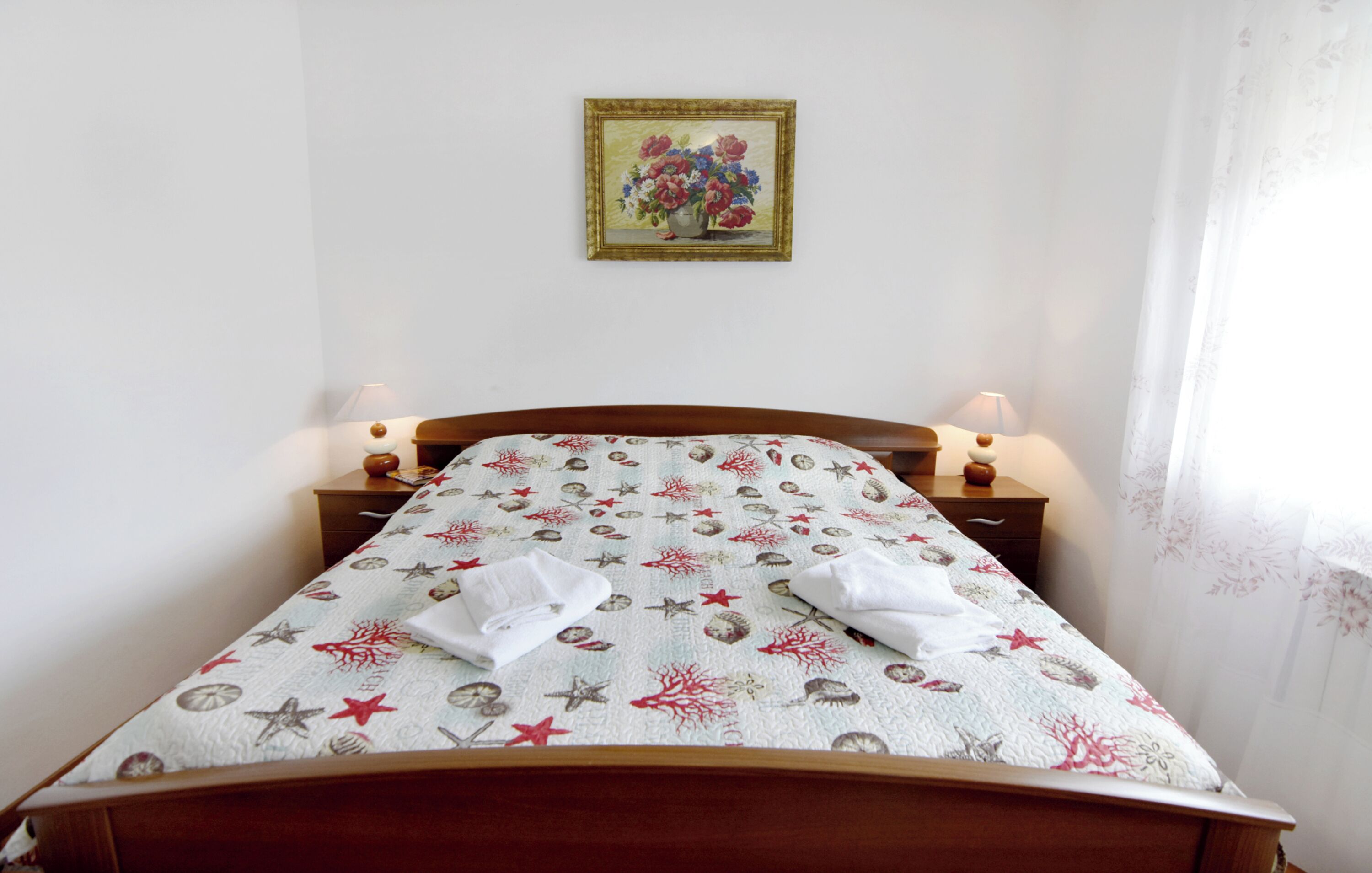 A neatly made bed with a patterned bedspread, two bedside lamps, and a framed floral painting on the wall.