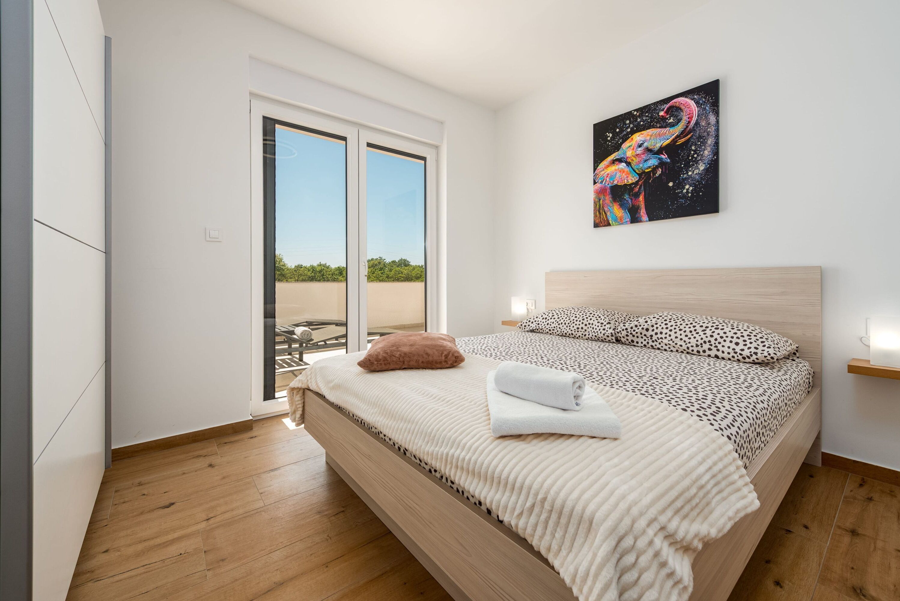 A bedroom with a large bed, a sliding glass door to a balcony, and a colorful elephant painting on the wall.