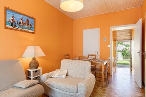 Ein Wohnzimmer mit einer Couch, einem Sessel, einem Tisch und einer Lampe. An der Wand hängt ein Gemälde, und eine Tür führt in einen anderen Raum.