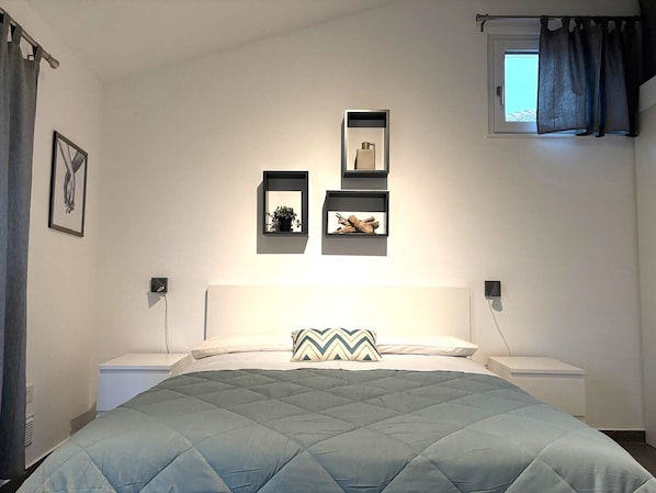 A bedroom with a bed, bedside tables, a wall-mounted shelf with decorative items, a window with curtains, and a framed picture on the wall. - HOUSEHOLD AT THE LAKE (Lavena Ponte Tresa)
