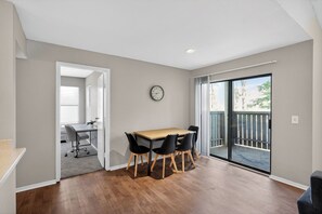 A room with a wooden dining table, four chairs, a clock on the wall, and a sliding glass door leading to a balcony.