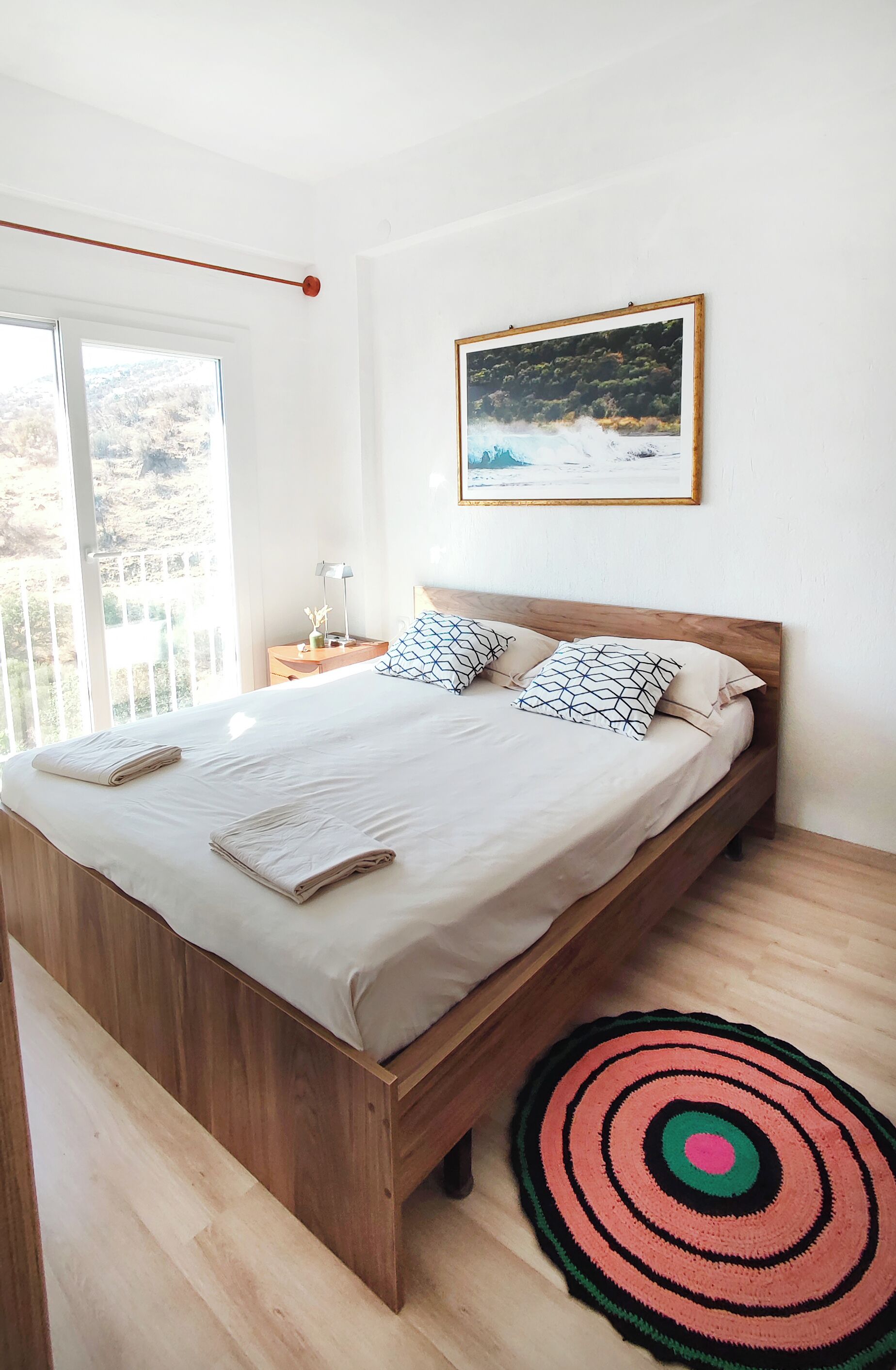 A bedroom with a wooden bed, a large window, a framed picture on the wall, and a circular rug.