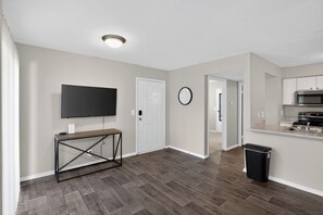 A modern living room with a flat-screen TV, a console table, a door, a clock, a trash can, and a kitchen area with a microwave.