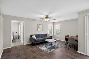 A room with a sofa, a round coffee table, a dining table with chairs, a ceiling fan, and a door leading to another room.