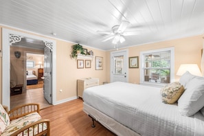 Une chambre à coucher comprenant un lit, un ventilateur de plafond, une porte, une fenêtre avec des stores et une table de chevet.
