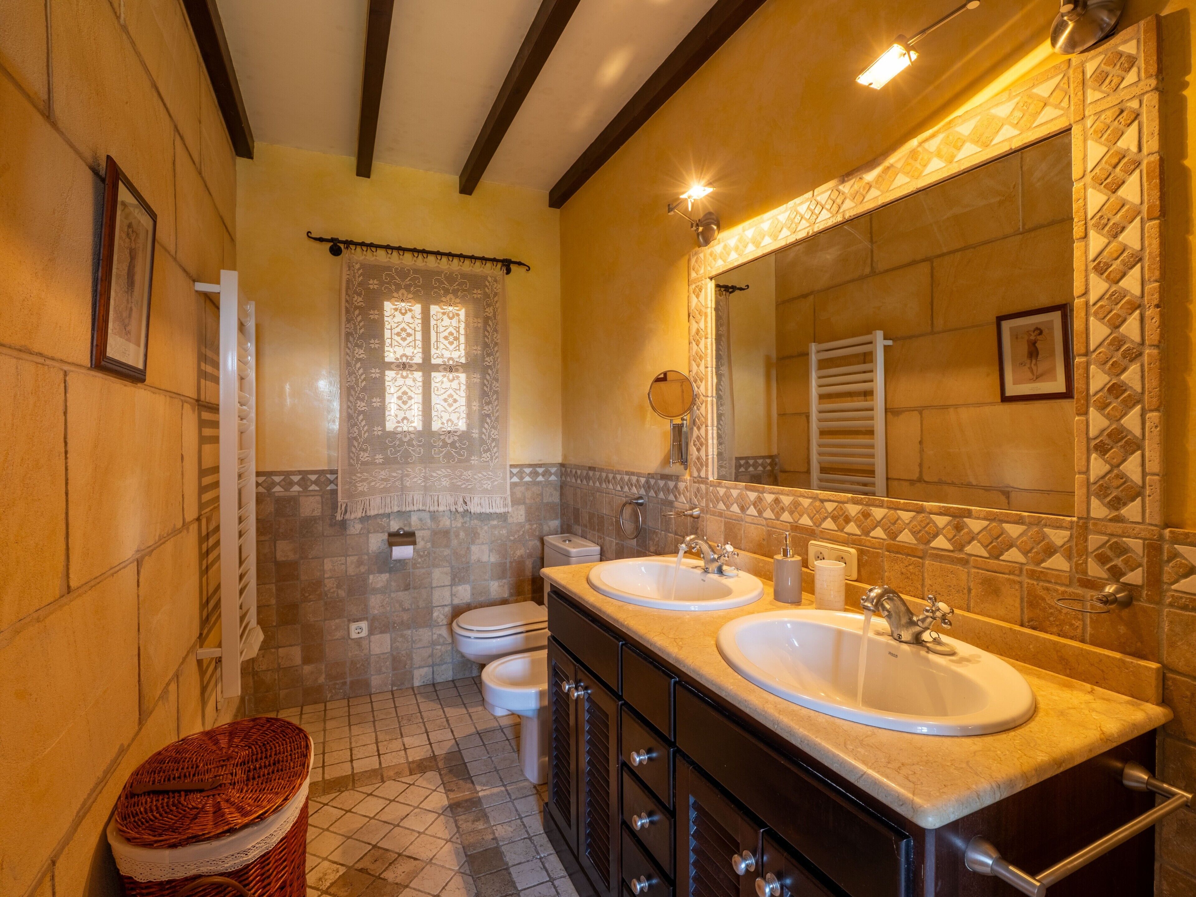 A bathroom with two sinks, a toilet, a window with lace curtains, and a mirror with a decorative frame.