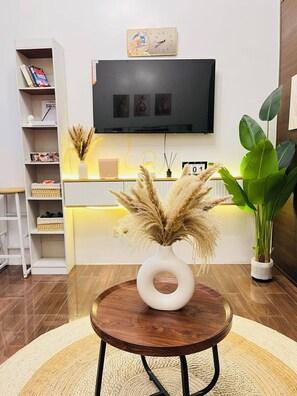 Ein Wohnzimmer mit einem runden Holztisch, einer weißen Vase mit Pampasgras, einem Bücherregal, einem an der Wand montierten Fernseher und einer großen Grünpflanze.