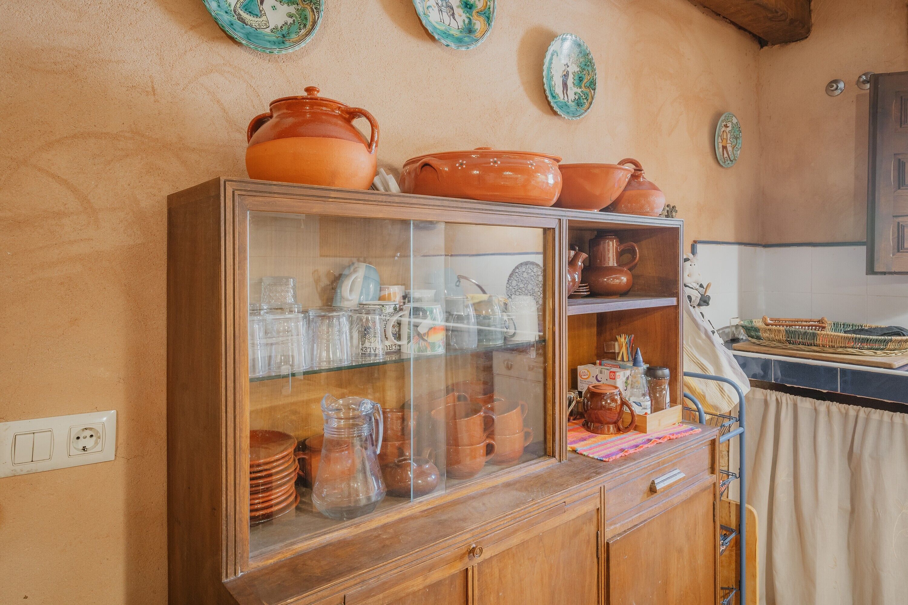 Una cocina con un armario de madera que contiene varios utensilios de cocina, una vitrina de vidrio y platos decorativos en la pared.