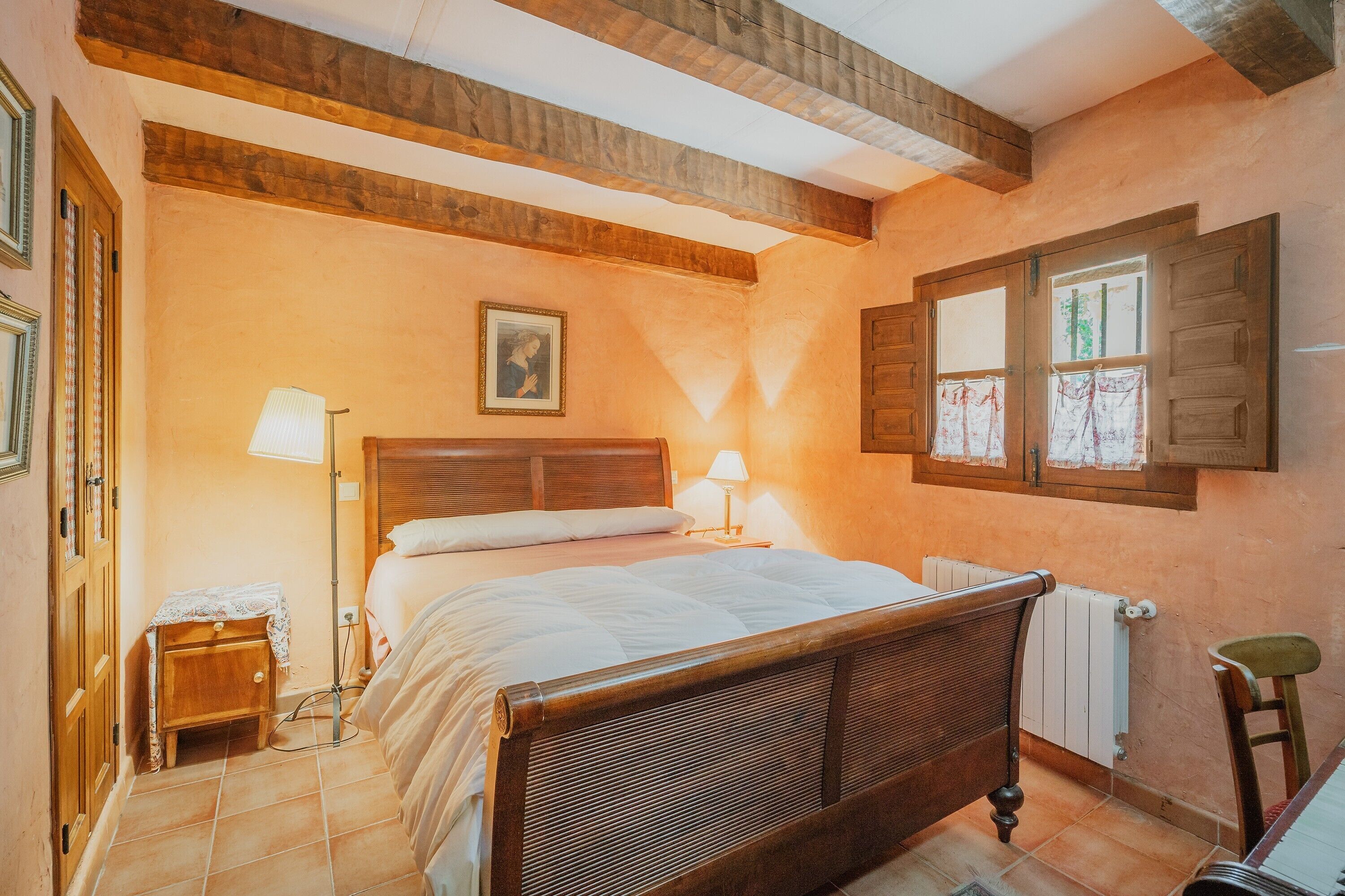 Un dormitorio con cama de madera, lámparas de noche, un radiador y una ventana con cortinas de encaje.