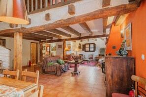 A rustic interior with exposed wooden beams, a dining area with wooden chairs, a sofa with a patterned throw, and a wooden table with a vase.