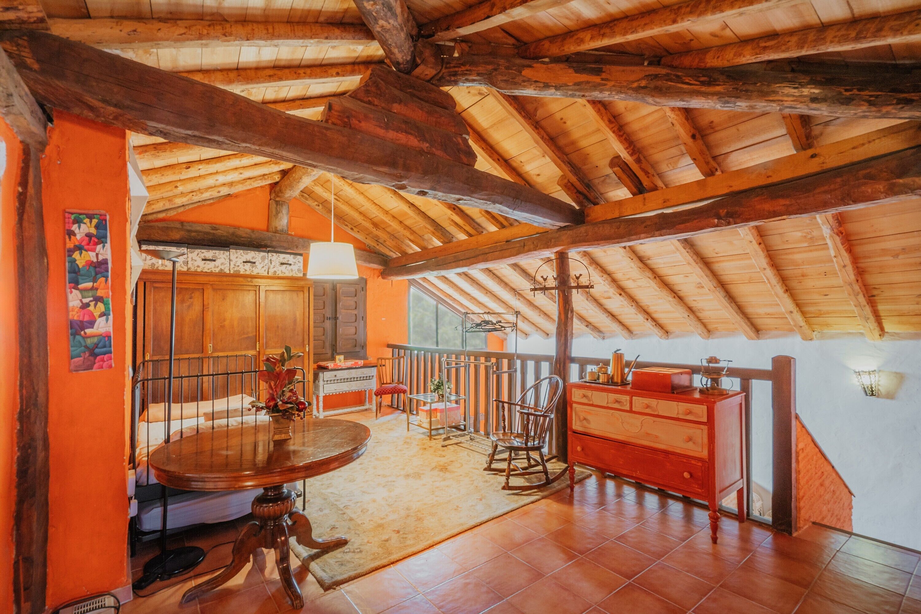 Interior rústico com teto de madeira, área de jantar com mesa redonda, cadeira de balanço e cômoda vermelha.