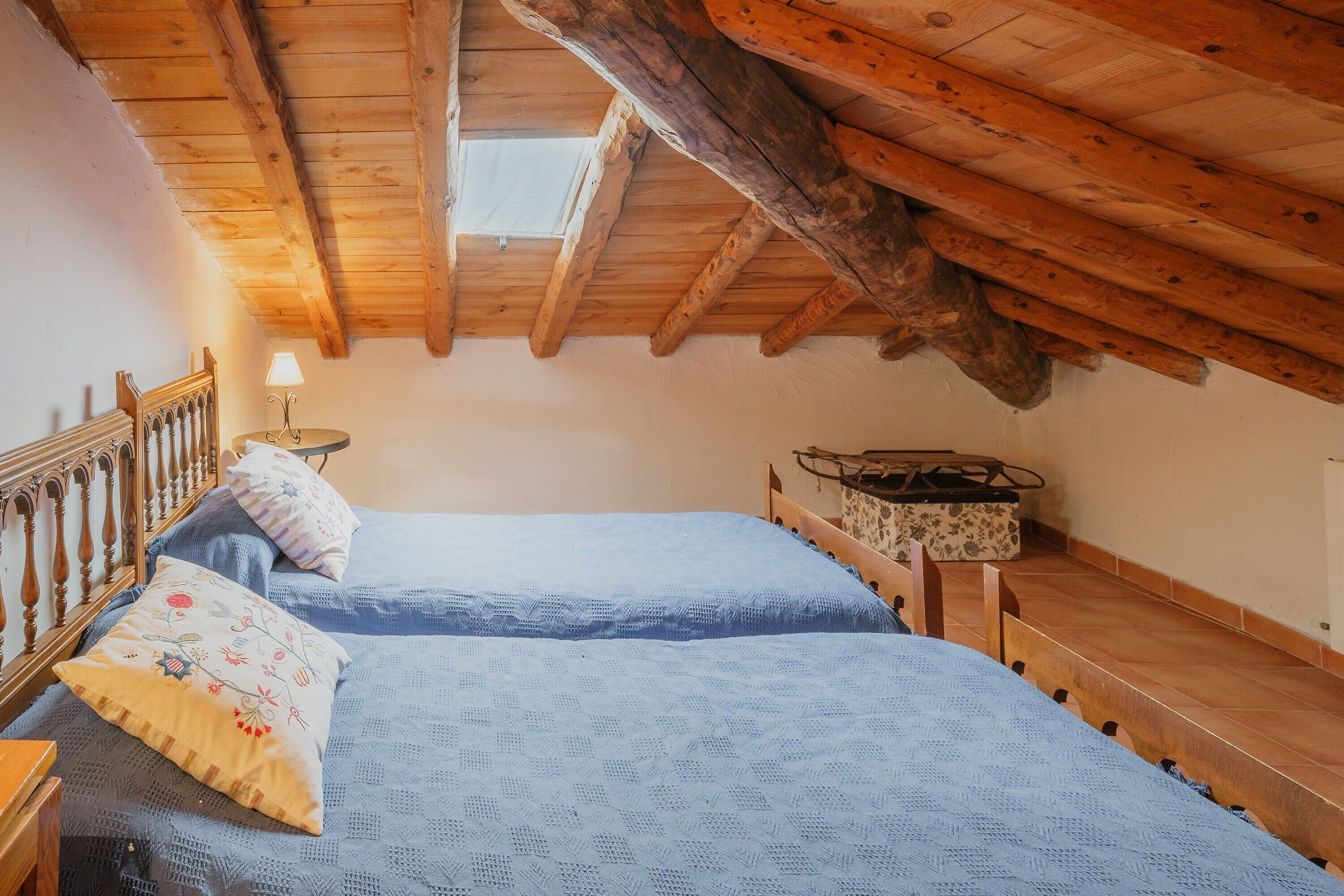 A bedroom with a sloped wooden ceiling, two beds with blue bedding, a nightstand, a lamp, and a wooden chair.