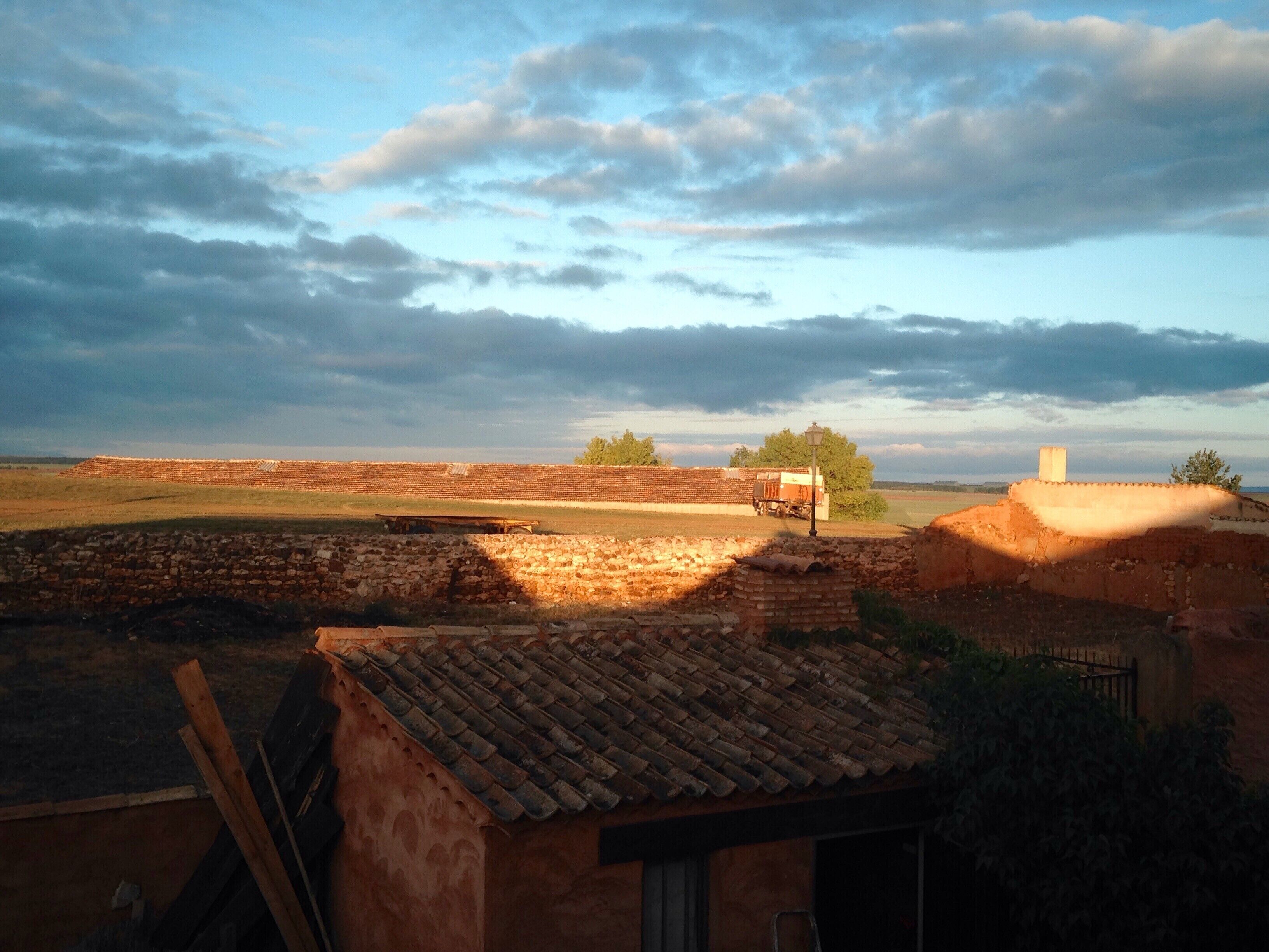 Paisaje rural con un muro de ladrillo, un pequeño edificio con techo de tejas y un camión a lo lejos.