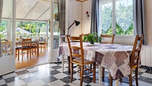 Un coin repas avec un sol à carreaux, des chaises en bois, une table recouverte d’une nappe à motifs et une vue sur l’extérieur, visible à travers une porte vitrée.
