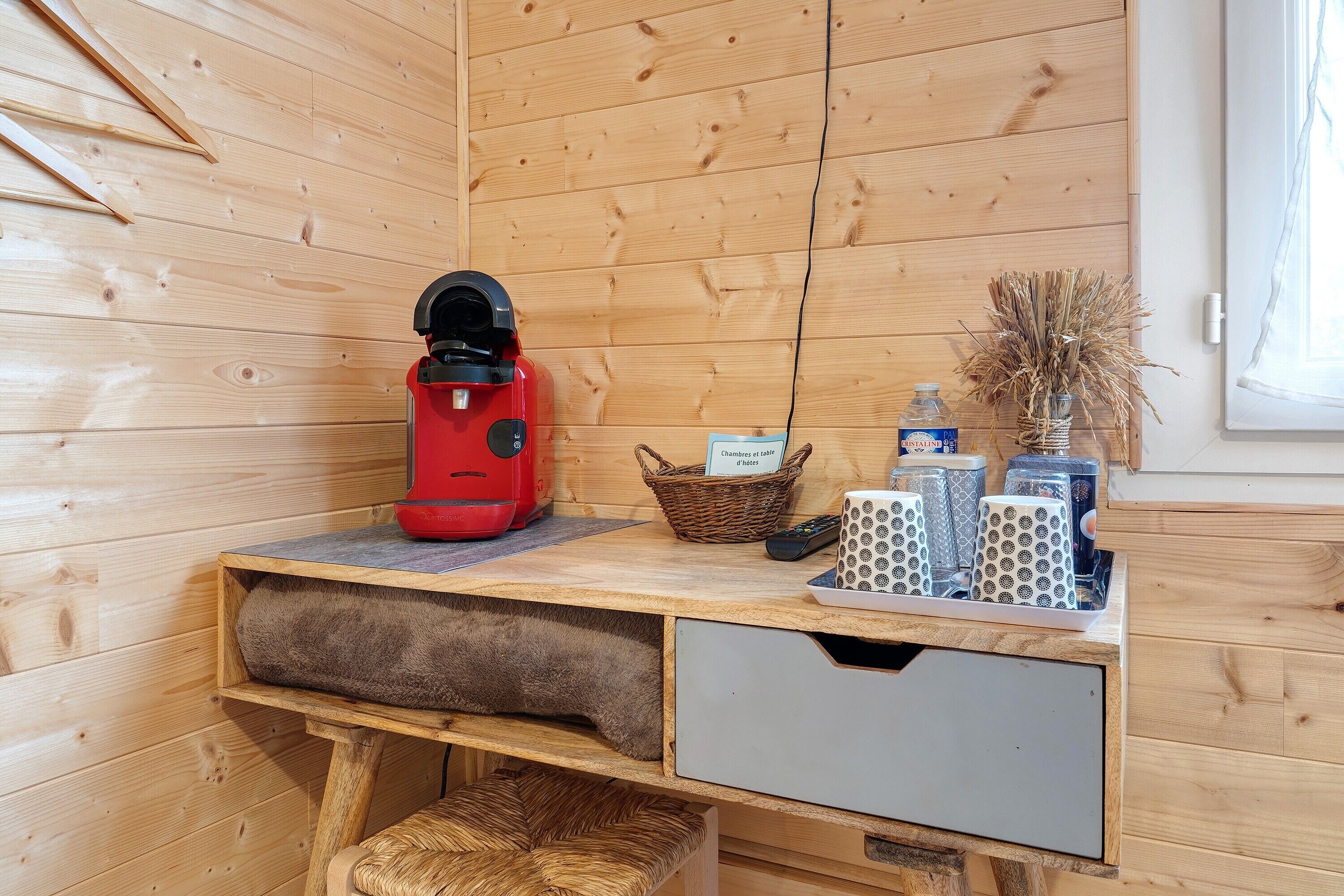 Ein Holzhütten-Interieur mit Schreibtisch, roter Kaffeemaschine, Korb und einer Vase mit Trockenpflanzen.