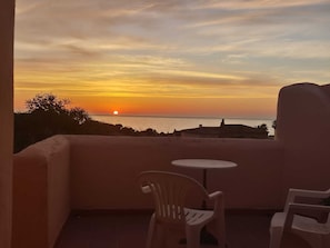 Ein Balkon mit Blick auf den Sonnenuntergang über dem Meer, zwei Plastikstühle und ein kleiner runder Tisch.