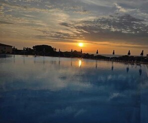 Ein Schwimmbecken spiegelt den Sonnenuntergang wider, im Hintergrund sind Gebäude und Bäume zu sehen.