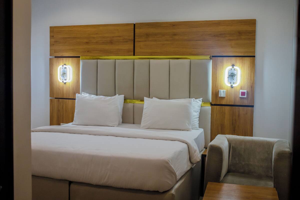 Ein Hotelzimmer mit einem großen Bett, zwei Nachttischen, einem Sessel und einem Holzkopfteil mit Wandleuchten.