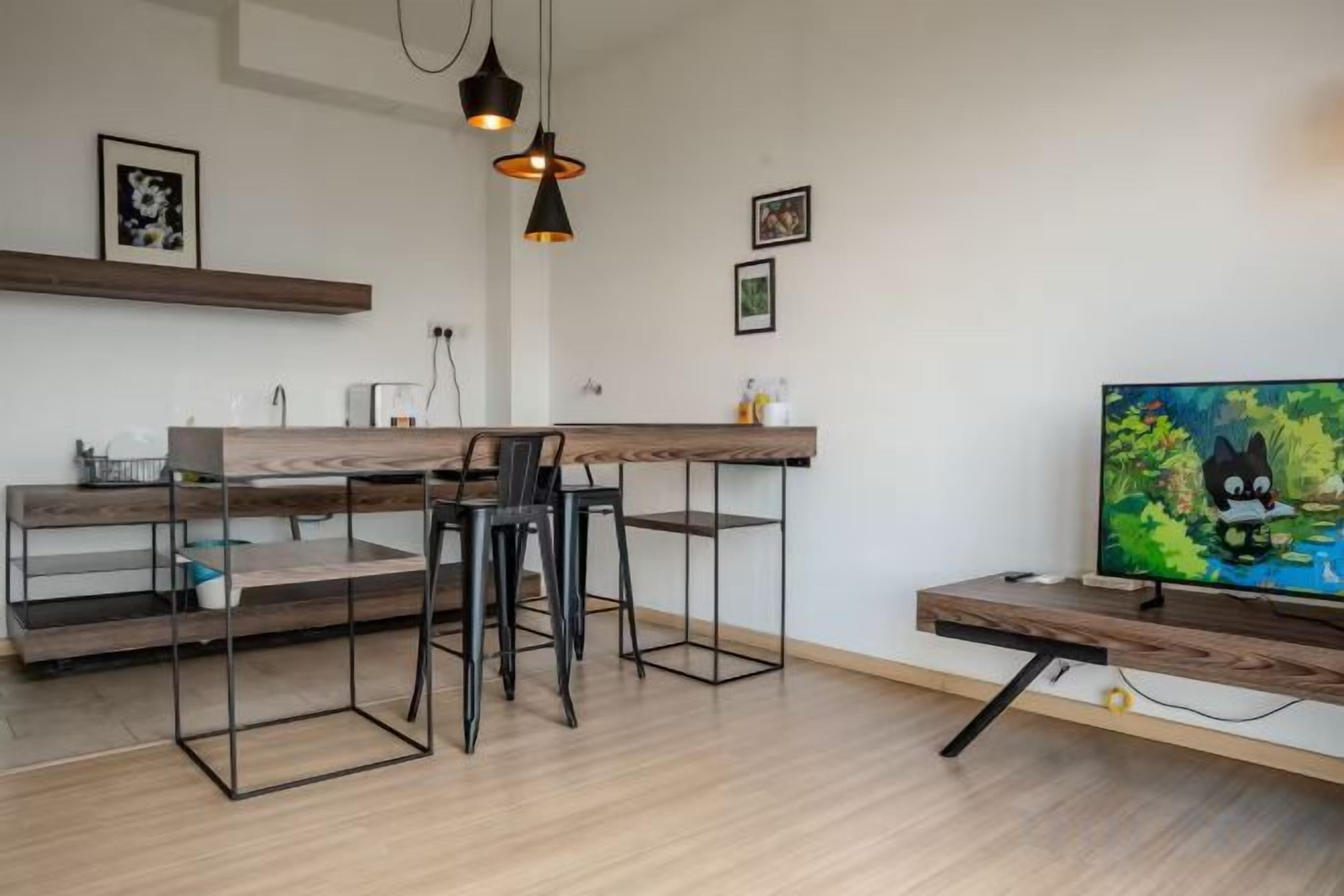 A modern kitchen with a wooden countertop, bar stools, pendant lighting, and a flat-screen TV on a wooden stand.