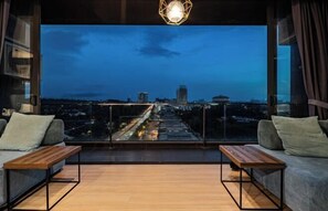 Una sala de estar moderna con una gran pantalla de televisión que muestra una vista urbana, dos sofás, una mesa de centro de madera y una lámpara colgante.