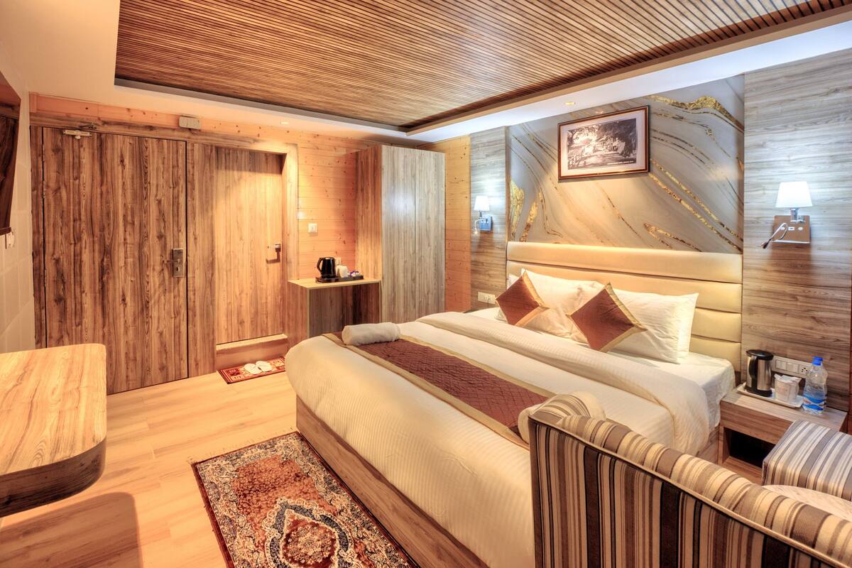 A hotel room with a wooden ceiling, a bed with multiple pillows, a bedside table with a lamp, and a small shelf with a coffee maker.