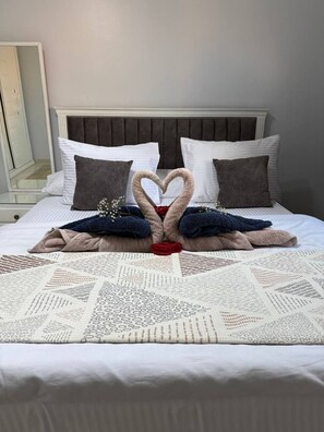 Una cama con una toalla dispuesta en forma de corazón, almohadas blancas y una colcha estampada.
