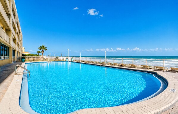 A clear blue pool with a wooden deck, a palm tree, and a view of the ocean. - Ocean View Condo | Beachfront Resort Escape (Daytona Beach)