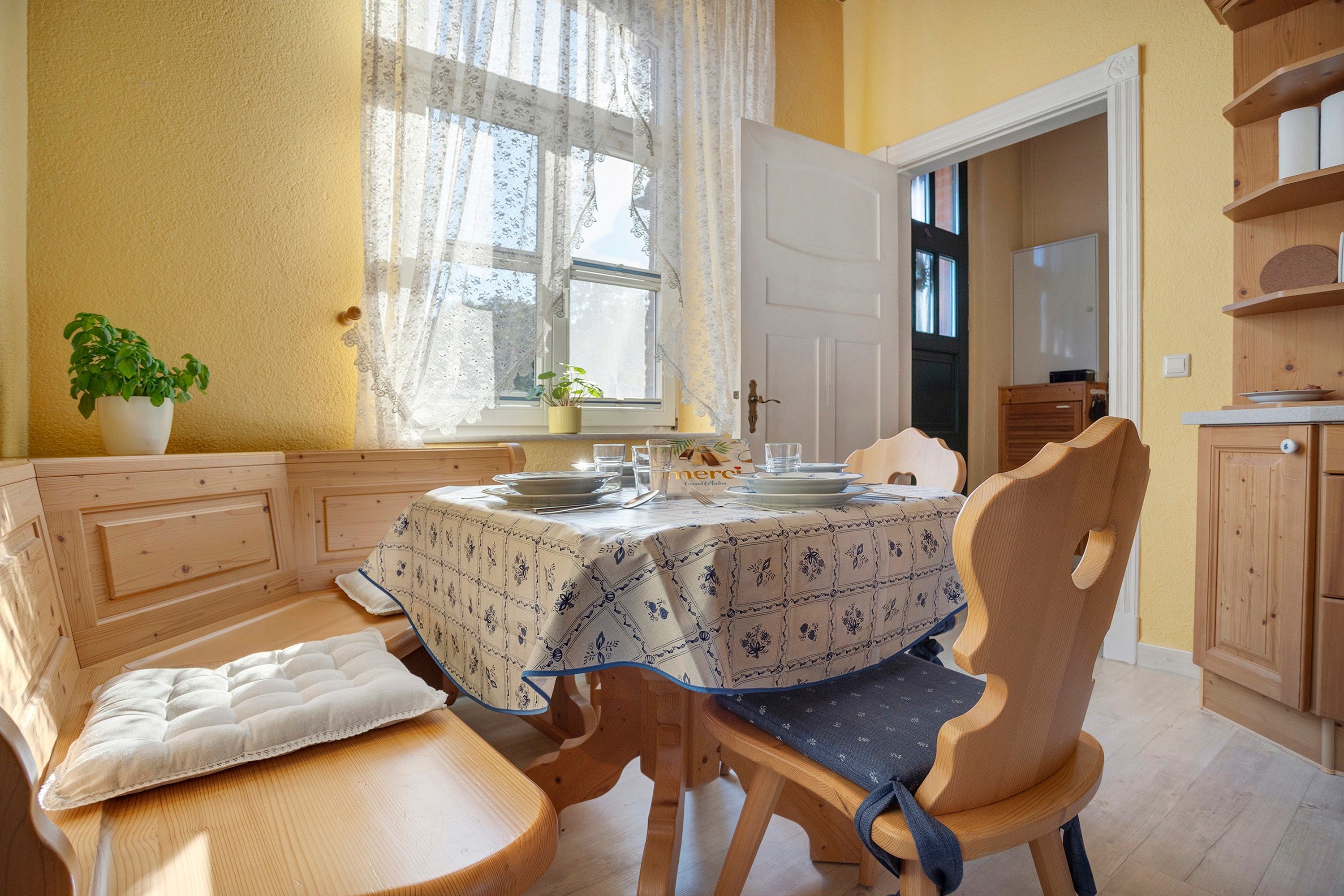 A dining area with a table set for two, a chair with a cushion, a potted plant, and a window with lace curtains.