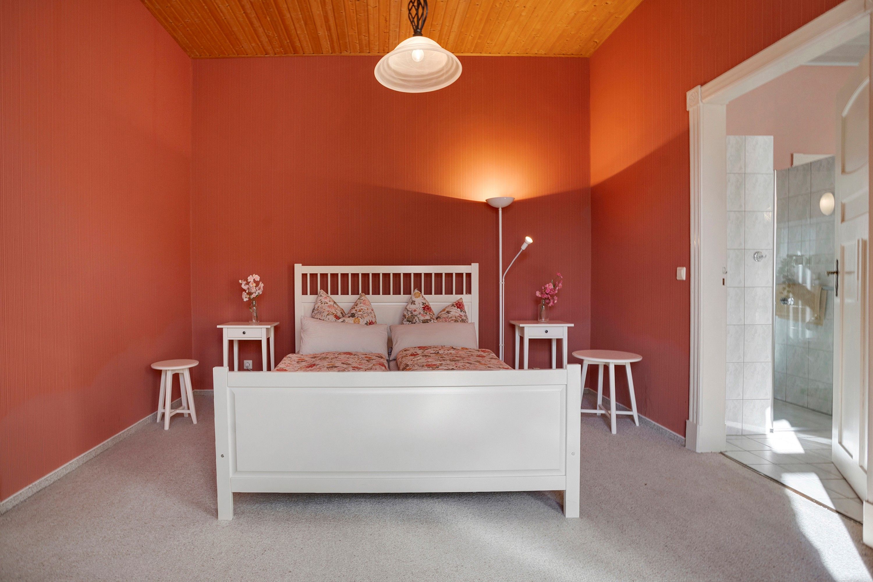 Une chambre à coucher avec un lit blanc, deux tables de chevet, une lampe sur pied et une porte donnant sur une salle de bain.