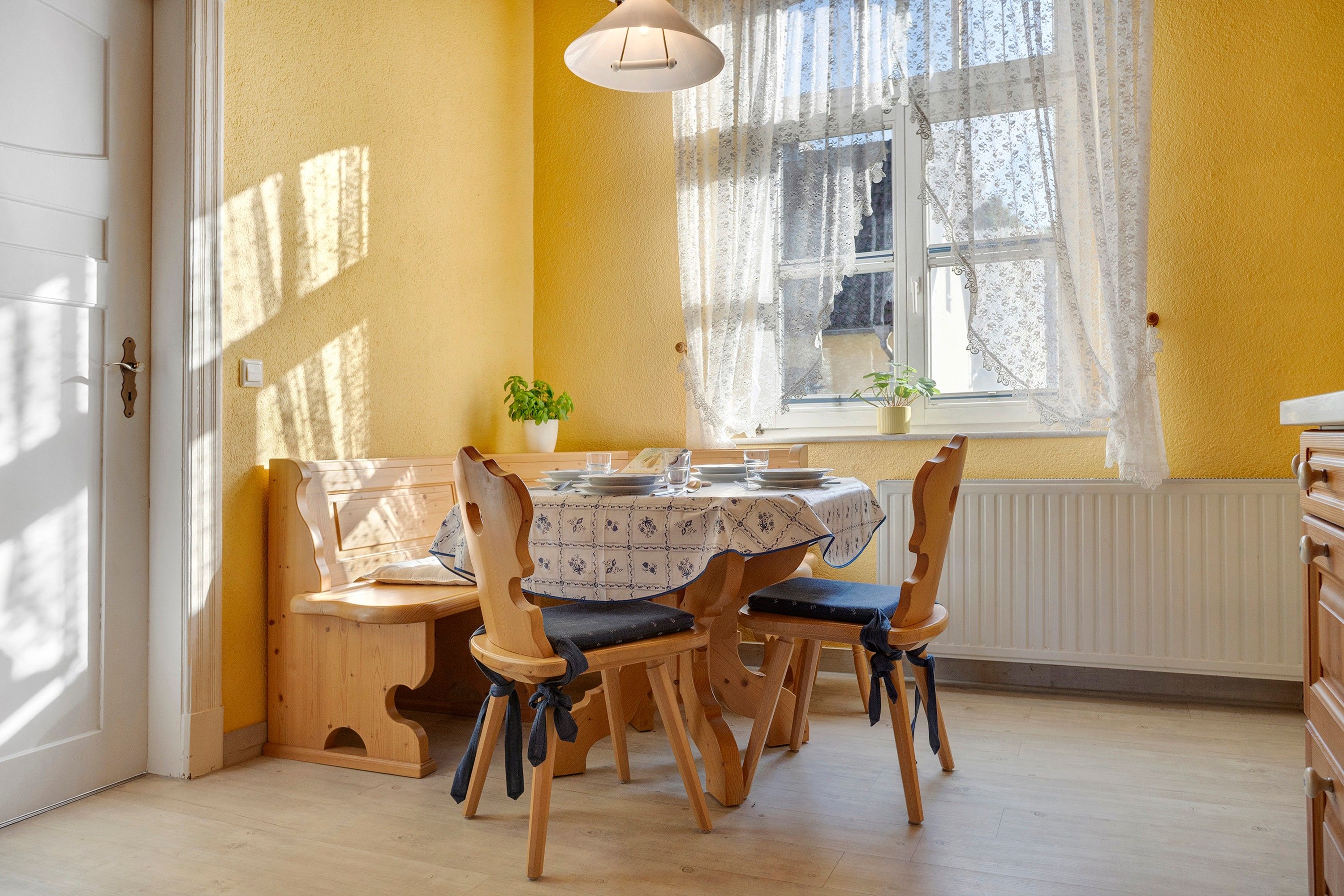 A dining area with a round table, wooden chairs, a bench, and a window with lace curtains.
