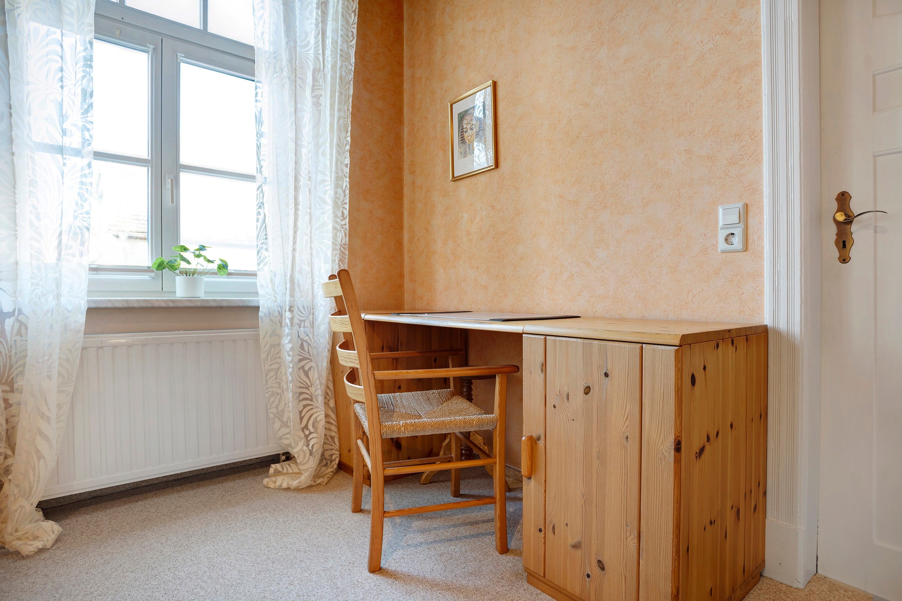 A room with a wooden desk, a chair, a radiator, a window with curtains, and a door with a light switch.