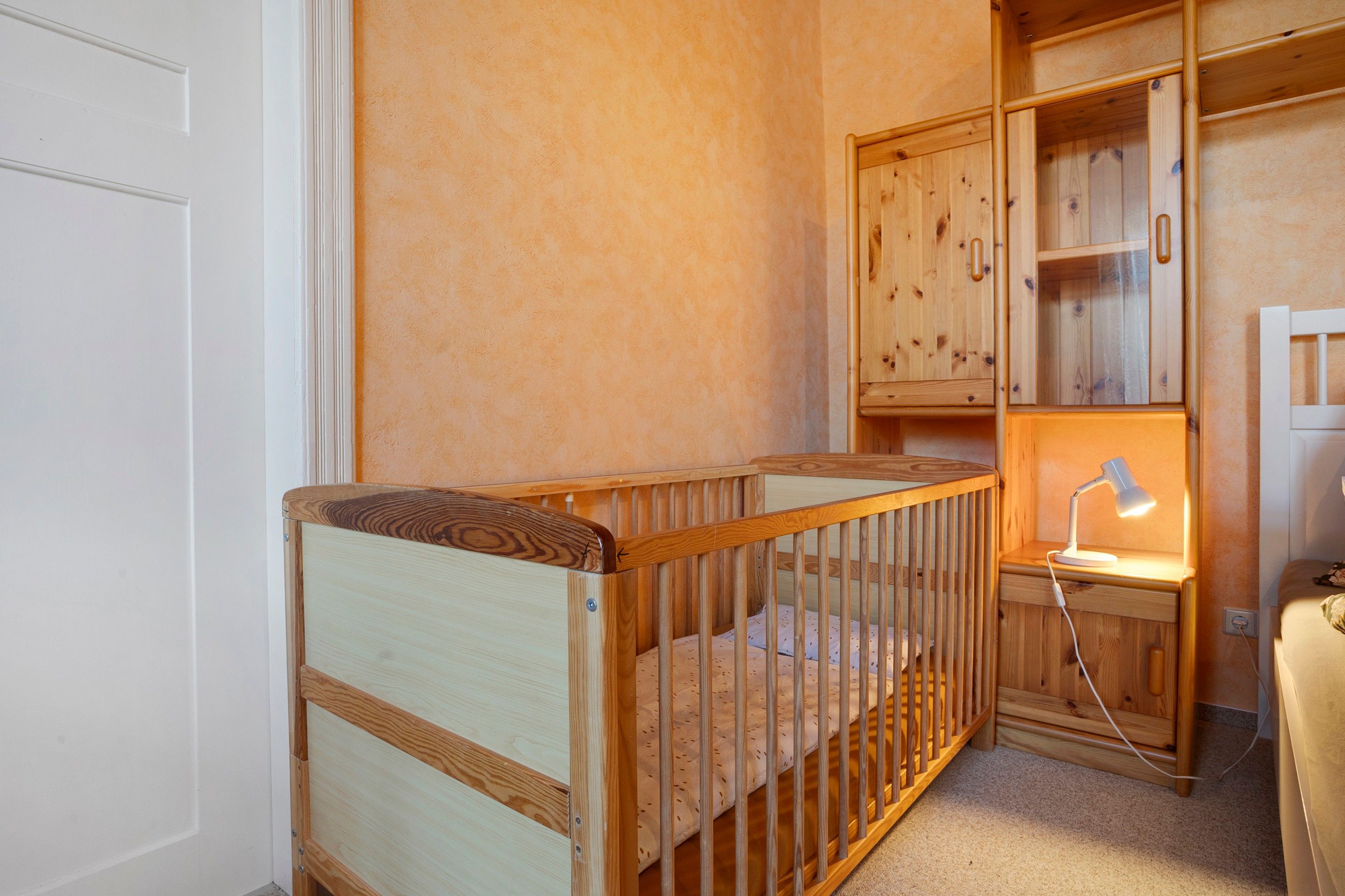 A wooden crib with a mattress, a nightstand with a lamp, and a wooden cabinet with drawers.