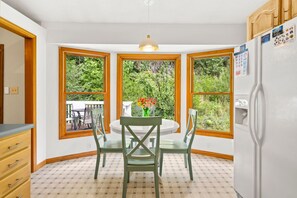 A kitchen with a dining table, chairs, a refrigerator, and large windows with a view of greenery.