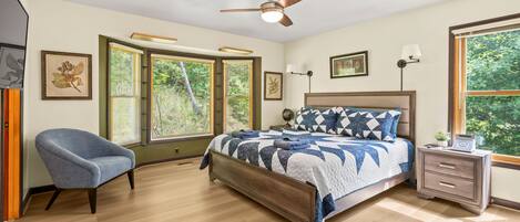 A bedroom with a bed, a chair, a nightstand, a ceiling fan, and large windows.