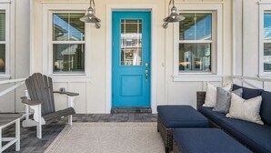 A porch with a blue door, two wall-mounted lamps, a gray Adirondack chair, a small table, a blue sofa with cushions, and a patterned rug. - Naples Luxury Cottage-Stylish 2BR in Premier Luxury Community (Naples)