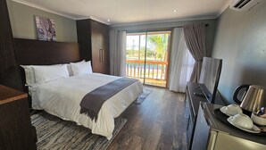 A hotel room with a large bed, a desk with a kettle, a TV, and a balcony with a view of greenery. - Zebra Hilltop Lodge (Mazunga)