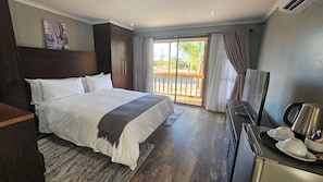 A hotel room with a large bed, a desk with a kettle, a TV, and a balcony with a view of greenery. - Zebra Hilltop Lodge (Mazunga)