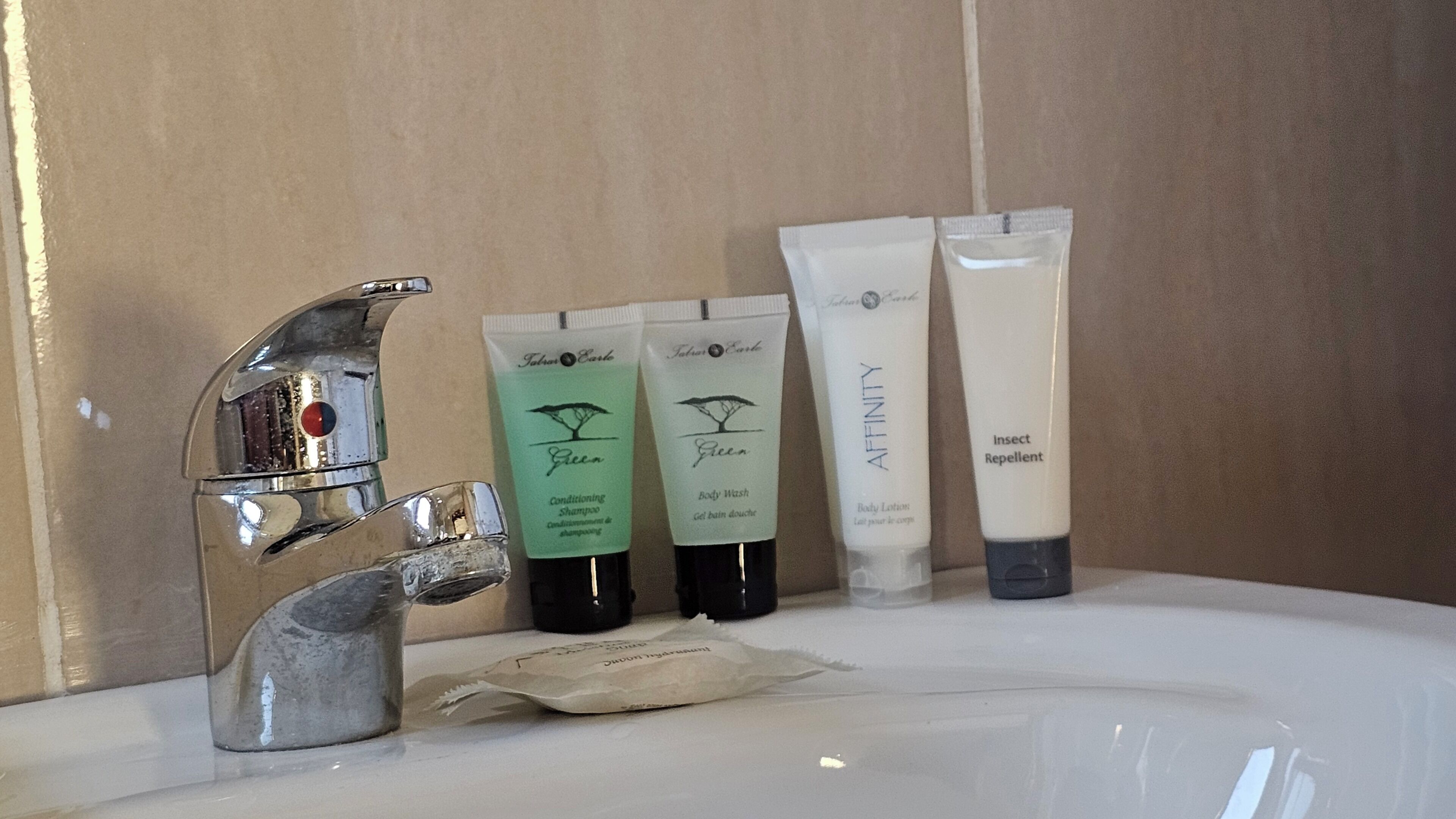 Un lavandino da bagno con un rubinetto cromato, tre flaconi di bagnoschiuma verdi e uno bianco, e un asciugamano color beige.