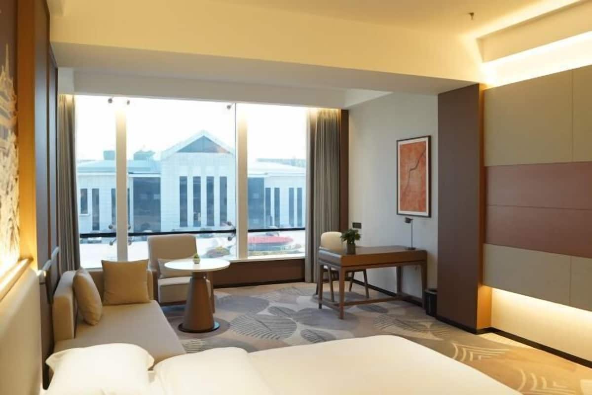 A hotel room with a bed, a sofa, a desk, a chair, and a large window with a view of a building.