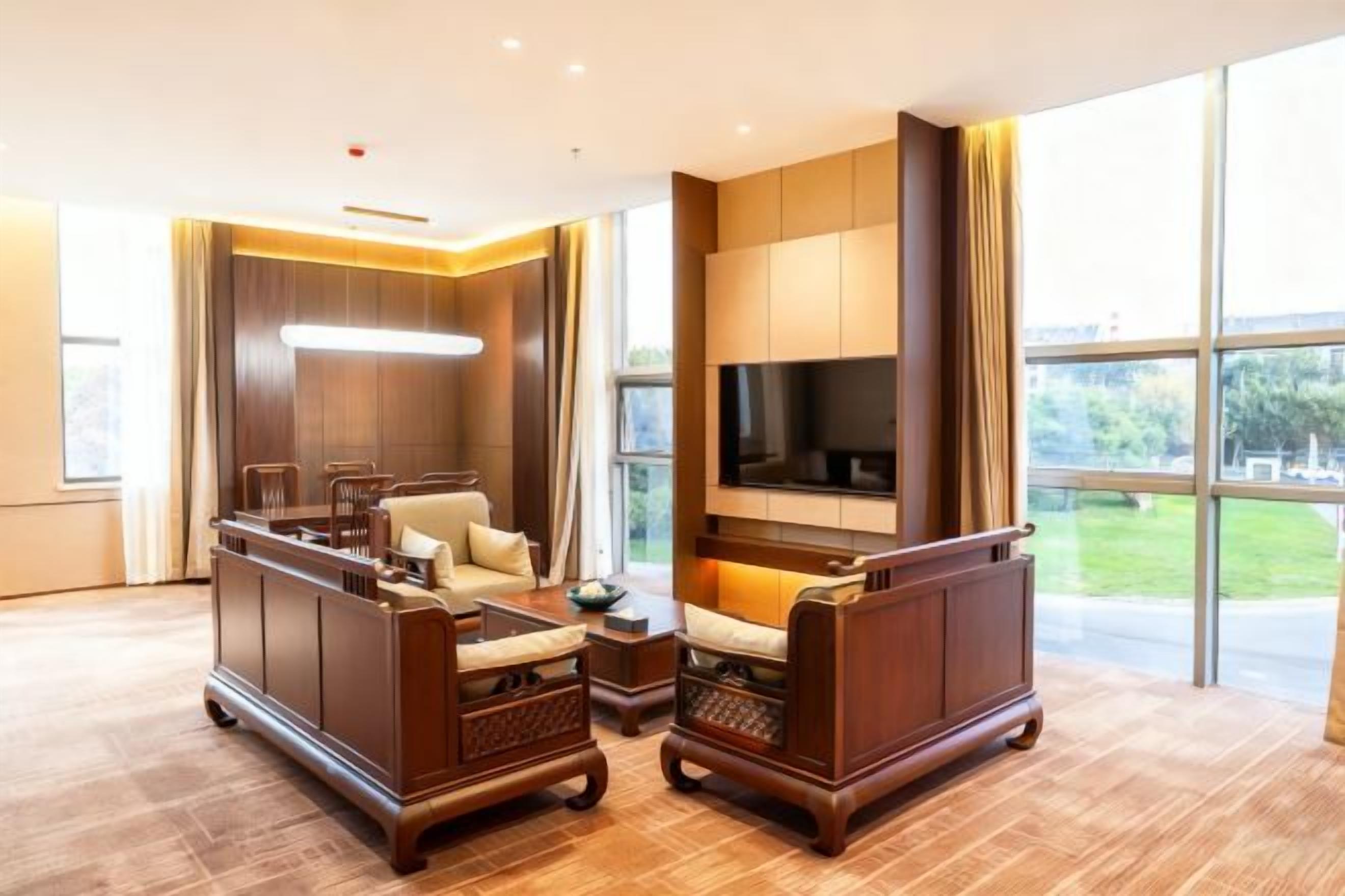 A spacious living room with a sofa set, a flat-screen TV mounted on the wall, and large windows offering a view of the outdoors.