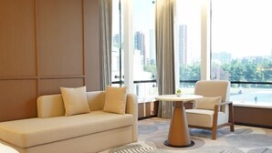Habitación de hotel con sofá, una silla, una mesita y vistas a un paisaje urbano a través de amplios ventanales.