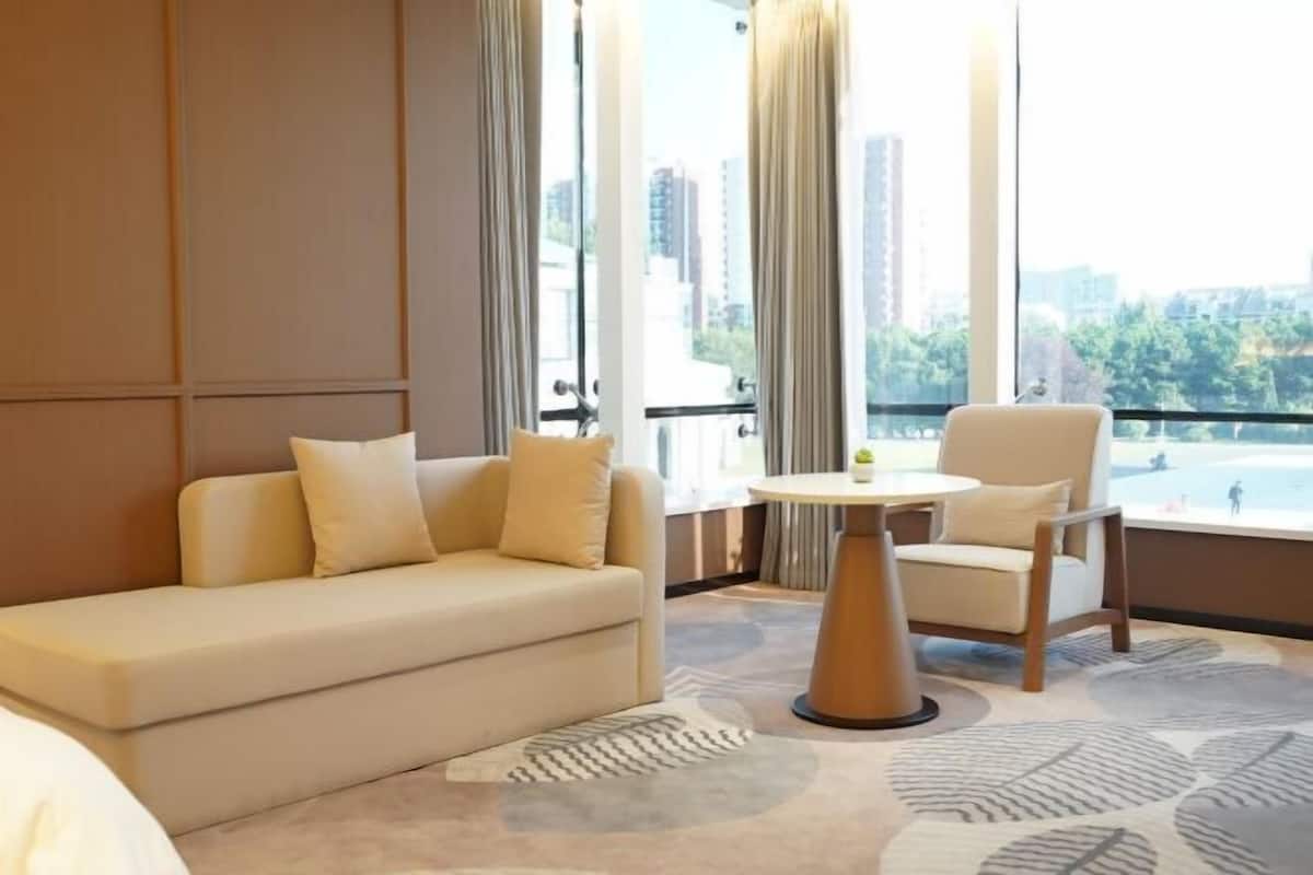 A hotel room with a sofa, a chair, a small table, and a view of a cityscape through large windows.