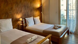 A hotel room with two beds, a wooden headboard, a nightstand, a lamp, a radiator, and a view of the outside through a window with curtains.