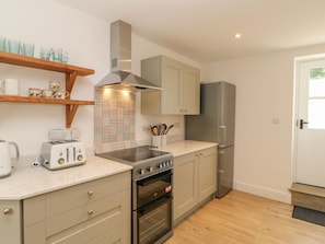 A compact kitchen with a built-in oven, a gas stove, a stainless steel refrigerator, and a wooden shelf with kitchenware. - Dairy Cottage (Kirkby Stephen)