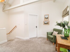A room with a white door, a green armchair, a wooden table, and two framed pictures on the wall. - Dairy Cottage (Kirkby Stephen)