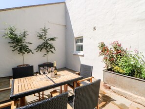 A patio with a wooden table and chairs, two potted trees, and a window. - Dairy Cottage (Kirkby Stephen)