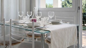 A dining area with a glass table, white chairs, and a view of greenery outside.